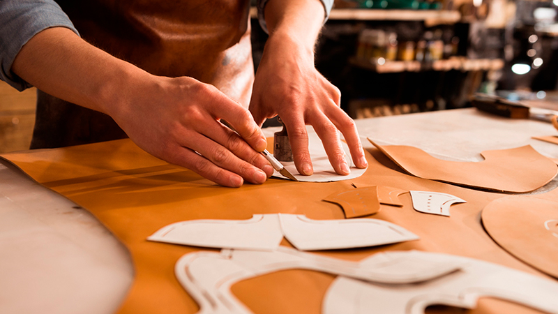 A shoemaker is hand-cutting leather into shaped pieces to form shoe upper